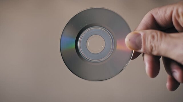 Top view of a male hand inspecting an old mini DVD disc with a reflective surface. The rainbow spectrum on the plastic highlights retro technology and data storage.