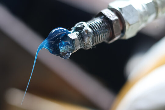 A macro close-up view of thick blue synthetic lubricant grease oozing and dripping from the coupler nozzle tip of a high-pressure grease gun inside an industrial workshop.