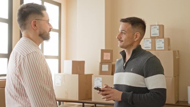 Two men exchanging a package as one hands it over and the other checks a smartphone, visible hands and boxes on shelves in a small business building; trust teamwork efficiency.