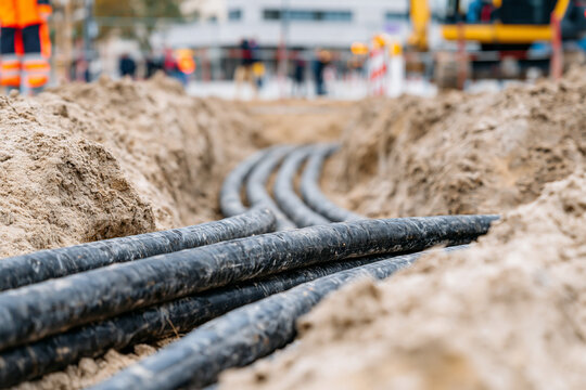 Thick industrial power cables laid in a trench on a construction site, surrounded by sand and machinery, with workers in safety gear visible in the background