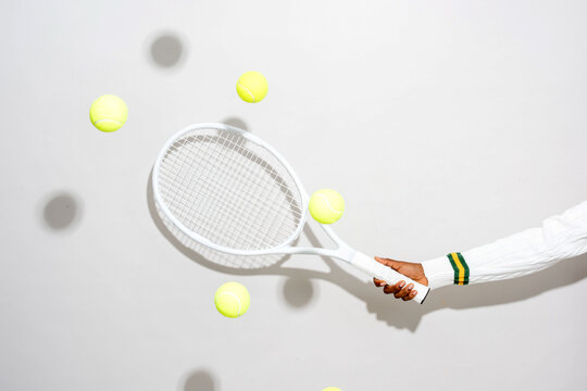 Hand holding a tennis racket with yellow balls mid-air on white.