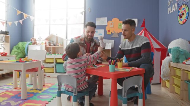Boy high fives with hand at red play table with colored pencils and toys, interacting with two men in a preschool play area inside a building; playful learning joy.