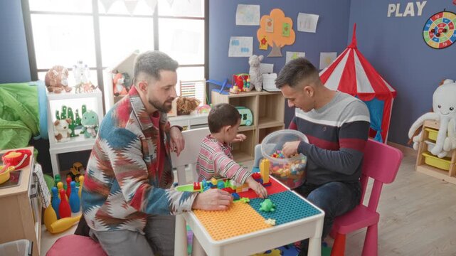 Men and child play with colorful plastic blocks as one man hands a block to the boy at a playroom building; family bonding joy.