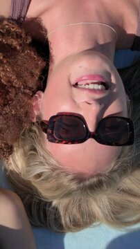 Vertical video: Smiling blonde woman lying on towel in sun, reacting to friend entering, mauve top