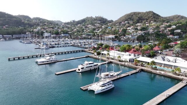 Aerial drone panning shot following luxury catamarans and sailing yachts lined up neatly at the harbor docks near the historic capital town center of Saint George's, Grenada