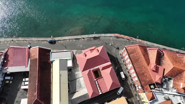Top down drone view looking down at colonial red-roofed waterfront warehouses and shallow emerald harbor channels along the coastal edge of Saint George's town, Grenada