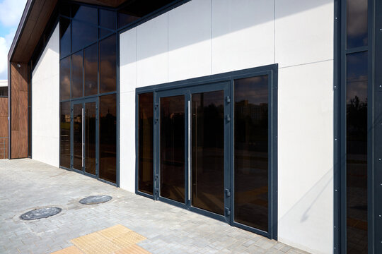modern building exterior features glass entrance doors with dark tint. white facade panels contrast with black metal frames. paved walkway includes manhole covers and tactile paving for accessibility