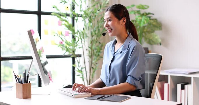 Friendly young asian woman working as a customer service representative, wearing a headset and smiling while providing support, assistance, and taking notes in a busy call center environment
