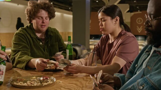 Diverse friends sitting at bowling alley table, arguing, counting cash and checking receipt to split bill for food and drinks. Peer-to-peer payment apps, budgeting, or shared expenses