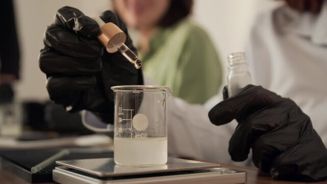 Young adult Caucasian woman in black gloves dripping essential oil from pipette into beaker on digital scale in slow motion. Clip is ideal for organic cosmetics ads