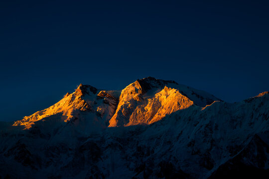 View of the snow-capped peak of Nanga Parbat glowing with golden light at sunset against a dark sky in Fairy Meadows, Gilgit, Pakistan.