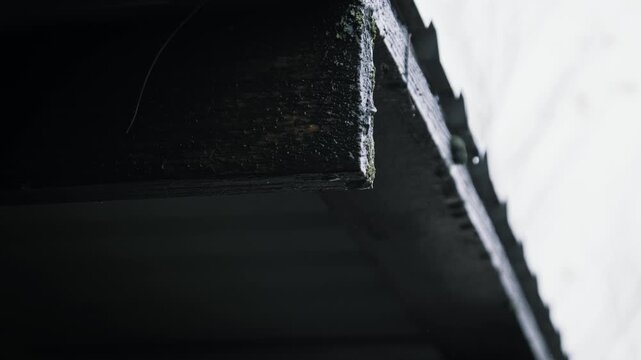 Clear rainwater droplets fall rhythmically from the corner of a wet dark wooden roof eave during a downpour. The weathered texture of the timber highlights the gloomy atmosphere.