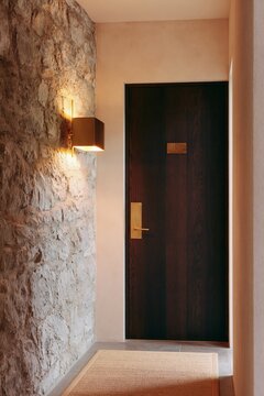 Boutique hotel hallway featuring rough stone walls, wooden door with gold handle, and wall-mounted light fixture illuminating the space