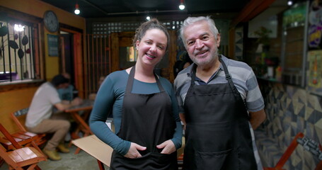 Obraz premium Young woman and senior man small business workers standing together inside café wearing aprons smiling at camera expressing teamwork pride and daily hospitality work