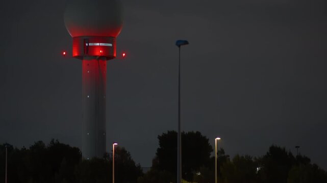 Airport surveillance radar tower glowing red at night with warning beacon lights active, street lamps and tree silhouettes visible in foreground against dark overcast sky