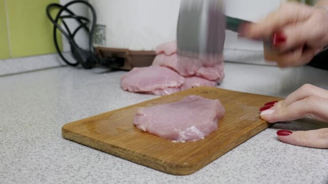 Female hands use a metal meat mallet to tenderize a raw pork steak on a wooden cutting board. The close-up shot focuses on the preparation process in a home kitchen setting.