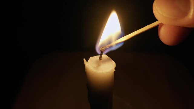Hand lights a single candle wick with a burning matchstick in a dark room. The flame flickers, casting a warm, soft glow against the black background as the camera pulls away.