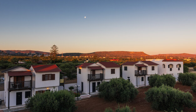 Scenic sunset landscape in Crete with white holiday villas, green trees and mountain ridges in the background during a golden hour. Kato Gouves, Greece island