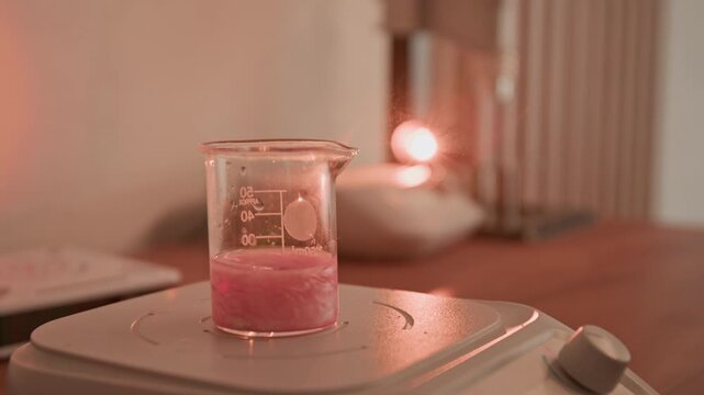 Gloved hand dropping solid ingredient into beaker with pink liquid on magnetic stirrer during perfumer master class. Clip is ideal for marketing artisanal perfume making or cosmetics formulation