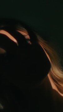 Woman with long blond hair shrouded in darkness sensually touching the skin of her arm as she turns