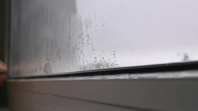 Water droplets from heavy condensation run down a foggy window pane, collecting on the white plastic frame. A close-up, static shot captures the details of moisture and dampness.