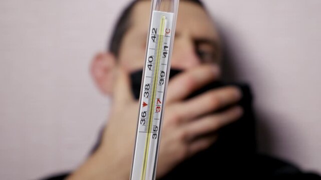 Man's hand holds glass mercury thermometer showing a high body temperature, indicating a fever. Camera focuses on the scale, with the sick, masked man blurred in the background.