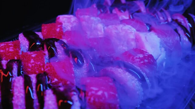 Assorted sushi rolls topped with roe and sauce sit on a platter under neon lights. Thick white dry ice smoke swirls around fresh Japanese food in pink and blue lighting.