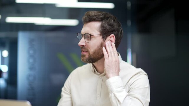 Close-up of businessman sitting in modern office suffering from ear pain. Male employee touching his ear with fingers, feeling discomfort from infection, otitis media, eczema, or other ear diseases.
