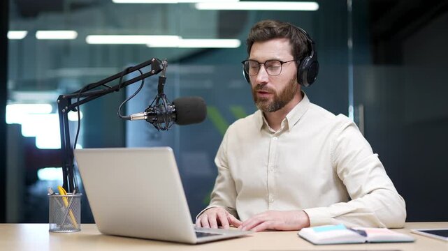 Confident businessman in wireless headphones recording podcast with microphone and laptop in modern office. Entrepreneur speaking during online webinar, virtual meeting, or live business presentation