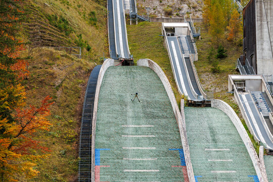 View of a ski jumper in mid-air over the green landing hill of the Planica Nordic Centre surrounded by autumn foliage in Planica, Kranj, Slovenia.