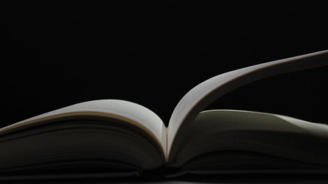 An open book rests in a dark room as the pages slowly turn and flutter, illuminated by a single light source against a solid black background. Concept of storytelling and knowledge.