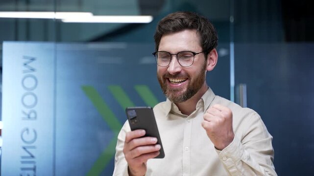 Close-up of happy businessman reading good news on smartphone in modern office. Smiling entrepreneur feeling pleased and satisfied after receiving positive message, enjoying successful moment at work