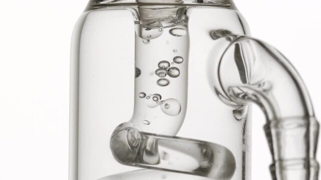 Hyper-close view of crystal clear liquid flowing through a glass tube, causing bubbles to condense and chaotically move inside it with a white backdrop | Allantoin solution beauty ingredient concept