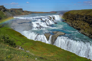 A wide river cascades over multiple waterfalls in a rugged landscape with green hills and a rainbow arching across the mist,Gullfoss,Iceland © SHUNJI ZHOU