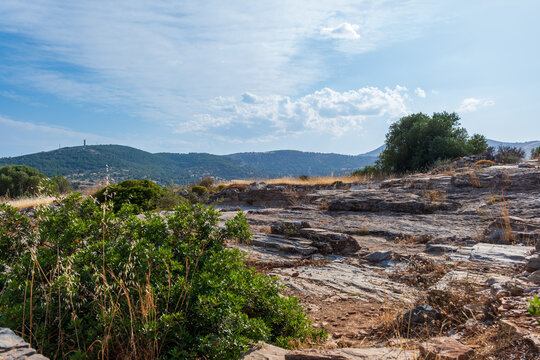 View of the Ancient Theatre of Thorikos archaeological site with rocky ruins and green bushes under a blue sky in Thoricus, Greece.