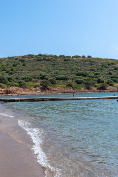 View of the Temple of Poseidon at Cape Sounio on a green hill overlooking a sandy beach and clear blue sea water in Sounion, Greece.