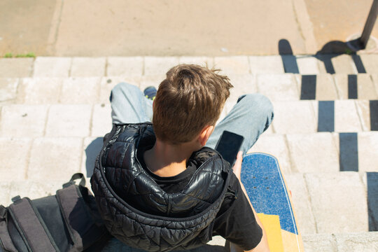Handsome teenager sitting with skateboard and texting on his phone
