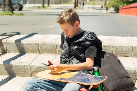 Handsome teenager sitting with skateboard and texting on his phone