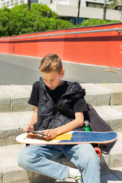 Handsome teenager sitting with skateboard and texting on his phone