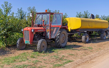 Obraz premium Old Red Tractor Pulling Yellow Water Tanker Cistern Trailer at Fruits Farm