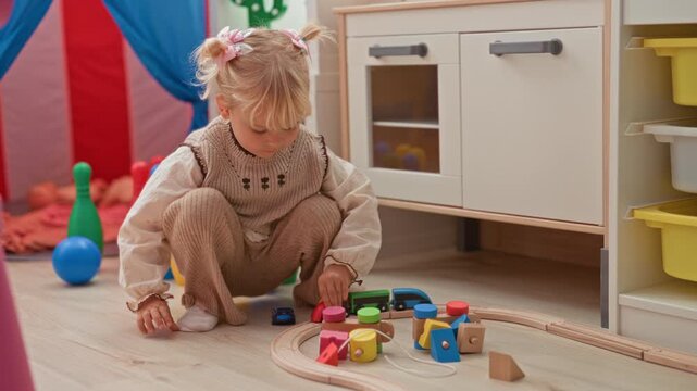 Little blonde girl kneeling, hands on wooden toy train and colorful blocks on floor in building; quiet concentration.