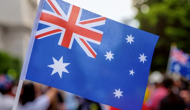 A small Australian flag waving proudly in a blurred outdoor setting, possibly during a national celebration or event.