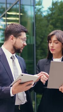 Two business colleagues, man and woman, talking and working on a laptop and tablet while walking outdoors near a modern office building. Slow motion