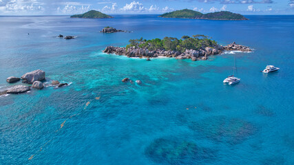 Stunning drone shot of Cocos Island, Seychelles, showing a luxury catamaran and boat anchored in clear turquoise water surrounded by granite rocks and coral reefs. © Nils