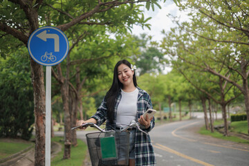 Naklejka premium Young Asian Woman Walking Bicycle on Cycle Path While Using Smartphone in Green Park