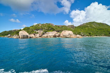 Naklejka premium Stunning coastal view of La Digue, Seychelles from a boat, featuring iconic granite rocks, lush green hills, and deep blue ocean waters under a cloudy blue sky