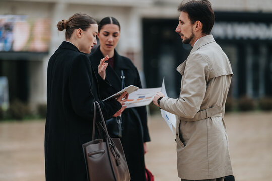 Three business professionals review charts and papers while standing outdoors. The colleagues discuss project details during an impromptu meeting in an urban setting.
