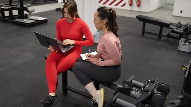 Two women pause gym training to review tasks on a laptop blending fitness and productivity Discussing projects planning schedules and balancing work with exercise