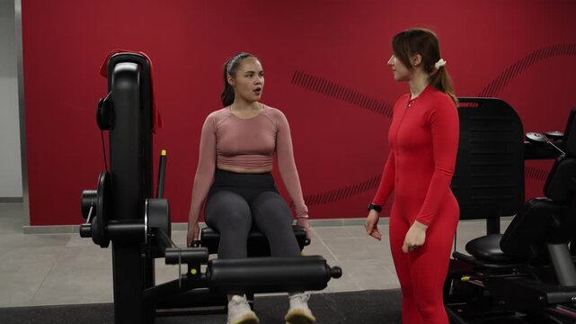 Personal trainer monitors client performing leg extension exercise on gym machine correcting technique and providing guidance for safe effective training
