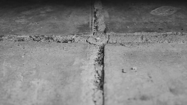 Monochrome low-angle photograph of cracked floor tile joints with dust, rough edges, shallow focus, and gritty construction surface.